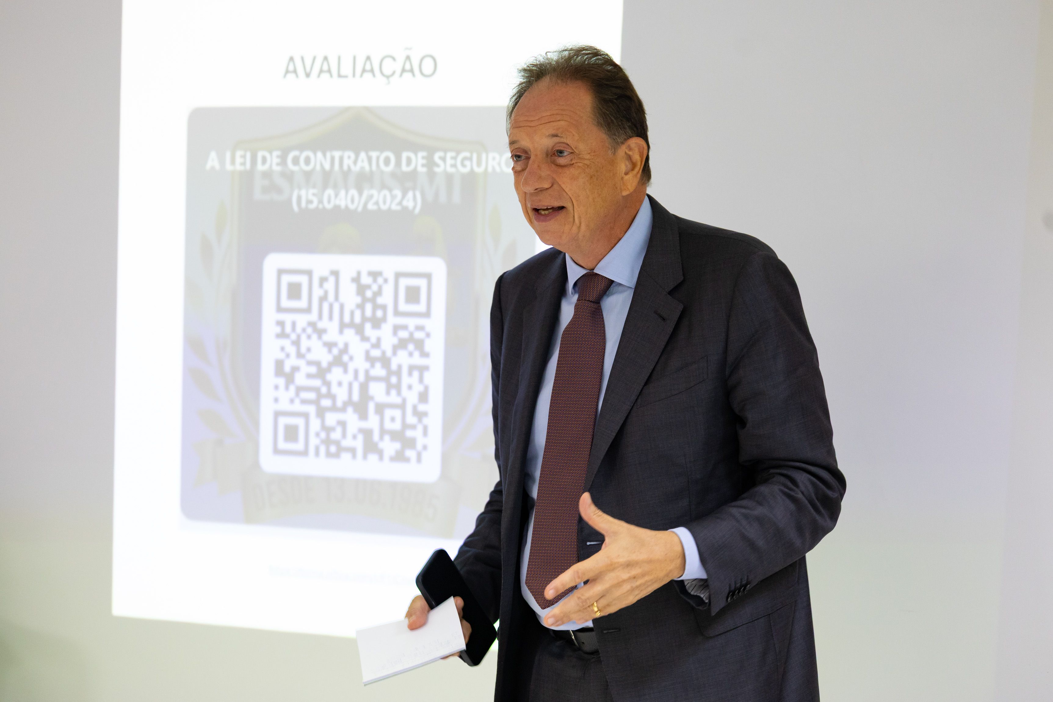 Homem em pé, usa terno escuro e gravata vermelha. Ele segura papel e celular, gesticulando enquanto fala diante de tela de projeção com QR Code e texto sobre avaliação do curso da Lei de Contrato de Seguro.